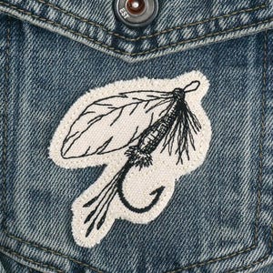 May include: A white embroidered patch of a fly fishing lure with black thread on a denim background.