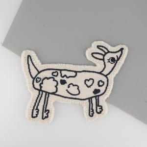 Könnte beinhalten: Gestickter Aufnäher mit einem Cartoon-Ziegen-Design. Die Ziege ist mit schwarzem Faden auf einem beigefarbenen Stoff umrandet, mit schwarzen Flecken und Herzdetails. Der Aufnäher hat einen Fransenrand.