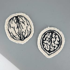 May include: Two off-white, oval-shaped embroidered coasters. Each coaster features a black embroidered walnut design. The coasters have a fringed edge and are set against a gray background. The design is a simple, yet elegant, representation of a walnut.
