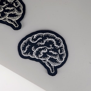 May include: Two navy blue embroidered brain patches with white stitching. The patches are in the shape of a human brain, with detailed lines representing the brain's folds and contours. The patches are likely for sewing or ironing onto fabric.