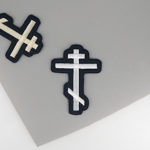 May include: Two embroidered Orthodox cross patches. The crosses are white with a black border. One cross is partially visible in the upper left corner. The other cross is centered and fully visible. The background is a light grey.