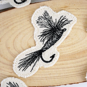 May include: A cream-coloured, embroidered patch in the shape of a fishing lure. The lure features a black hook and detailed black thread work, with a fluffy, textured appearance. The patch is likely for sewing or ironing onto fabric.