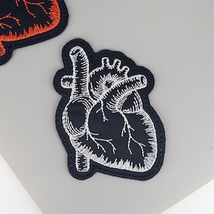 Peut inclure: Écusson brodé représentant un cœur anatomique détaillé en fil blanc sur fond bleu marine. Le patch a une bordure noire. L'image montre un gros plan du patch, soulignant la couture complexe et le contraste des couleurs.