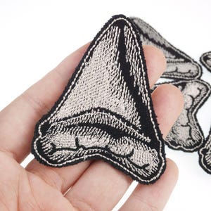 May include: Embroidered shark tooth patches in black and white. The patches have a detailed design with a black border and a white, textured interior. These patches are likely for decorative use, such as on clothing or accessories.
