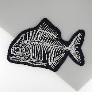 May include: A black embroidered patch featuring a white fish skeleton design. The patch has a textured, fuzzy border. The detailed skeleton design is set against a black background. Suitable for clothing or accessories.