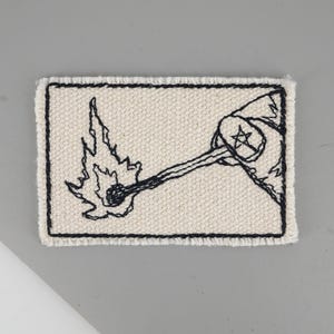 Op de afbeelding: Geborduurde patch met een zwart-wit ontwerp van een aangestoken lucifer. De lucifer wordt vastgehouden door een hand, met een stersymbool op de mouw. De patch heeft een rechthoekige vorm met een crèmekleurige achtergrond en een zwarte rand.