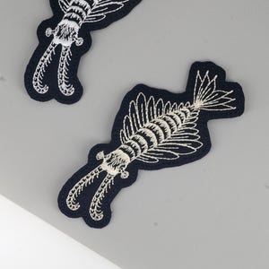 Puede incluir: Parches bordados con un diseño blanco detallado de un camarón mantis sobre un fondo azul marino. Los parches tienen un borde negro y se muestran sobre una superficie gris. El diseño es intrincado y muestra el cuerpo segmentado y los apéndices del camarón.