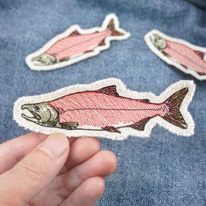 Può includere: Tre toppe di salmone ricamate su uno sfondo di denim blu. I salmoni sono rosa con dettagli marroni e verdi. Le toppe sono cucite sul denim.