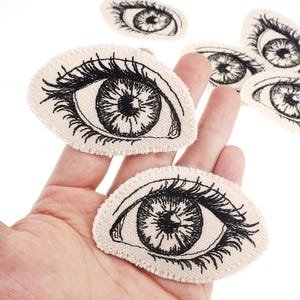 May include: Eye-shaped fabric patches with a detailed black and white eye illustration on each. The patches have a textured, off-white background. Suitable for sewing or crafting, these patches add a unique touch to projects.