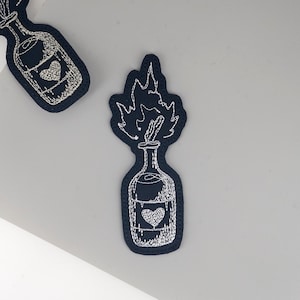 May include: Navy blue embroidered patches depicting a bottle with a heart and flames. The patches are detailed with white stitching. The bottle design includes a heart shape, and the flames are stylized.