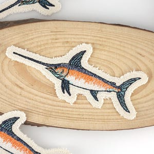 Può includere: Toppe ricamate con un design di pesce spada. Le toppe hanno un bordo color crema e sono posizionate su una superficie di legno. Il pesce spada presenta dettagli arancioni, blu e neri. Ideali per abbigliamento o accessori.