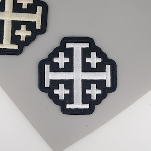 Puede incluir: Dos parches bordados con el diseño de la Cruz de Jerusalén. Los parches tienen un fondo negro con una cruz blanca y cuatro cruces más pequeñas en cada cuadrante. Los parches son probablemente para ropa o accesorios.