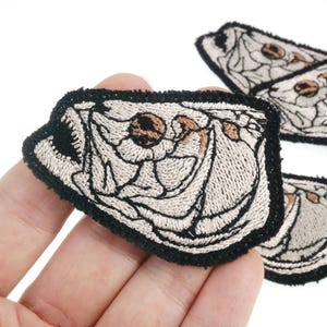 May include: Embroidered patches with a detailed design of a stylized animal head with open mouth, in beige, brown, and black. The patches have a black border, suitable for clothing or accessories.