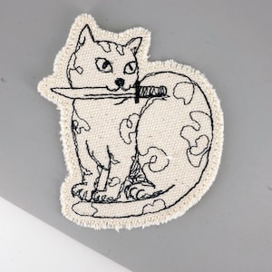 Puede incluir: Parche bordado con un gato sosteniendo un cuchillo en la boca. El gato es de color crema con detalles bordados en negro, incluyendo el contorno, rasgos faciales y remolinos decorativos. El parche tiene un borde deshilachado.