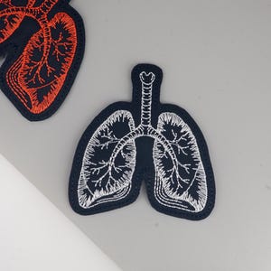 Könnte beinhalten: Gestickte Aufnäher in Form von Lungen. Einer ist schwarz mit weißen Nähten, der andere schwarz mit orangefarbenen Nähten. Die Aufnäher befinden sich auf einer grauen Oberfläche.