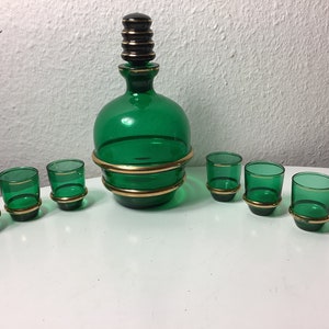 Puede incluir: Un juego de decantador de vidrio verde vintage con seis vasos de chupito a juego. El decantador tiene un tapón negro y bandas doradas. Los vasos de chupito también tienen detalles dorados. El juego se exhibe sobre una superficie blanca.