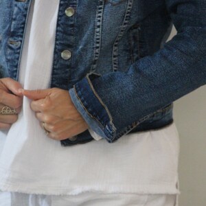 Puede incluir: Una persona lleva una chaqueta vaquera azul sobre una camiseta blanca. La chaqueta est&aacute; desabrochada y la persona est&aacute; ajustando el cuello.