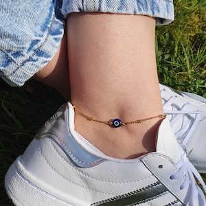 May include: A gold anklet with a blue evil eye charm. The anklet is worn on a person's ankle.