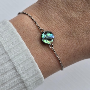 May include: Abalone shell bracelet with a silver chain. The round abalone shell has iridescent colors of blue, green, and purple. The bracelet is a delicate piece of jewelry.