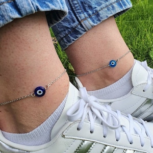 May include: A pair of silver-toned metal chain anklets, each featuring a single circular glass charm with a blue, white, and black evil eye design. The anklets are worn around the ankles above white socks and white sneakers with silver accents.
