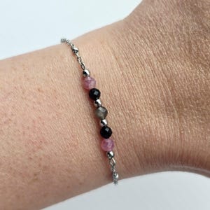 May include: A delicate bracelet featuring a silver chain with alternating pink, black, and gray gemstone beads. The bracelet is designed with small silver beads between the gemstones. The bracelet is shown on a wrist.