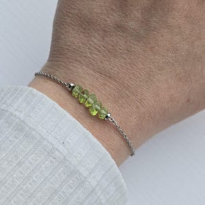 May include: A silver chain bracelet adorned with a line of light green peridot gemstones. The gemstones are cut and have a natural shape. The bracelet is worn on a wrist, against a white shirt. The gemstones are approximately 0.5 cm in size.