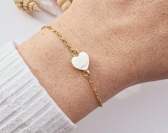 May include: A delicate gold chain bracelet with a white heart charm. The bracelet is on a person's wrist.
