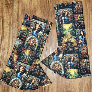 May include: A pair of yellow and black leg warmers with a stained glass pattern featuring characters from the Wizard of Oz. The pattern includes Dorothy, the Scarecrow, the Tin Man, the Cowardly Lion, and the Wicked Witch of the West.
