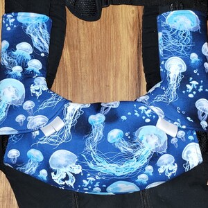 May include: Baby carrier with blue fabric featuring a jellyfish pattern. The fabric wraps around the carrier's straps and front panel. The jellyfish are white and light blue against a dark blue background.