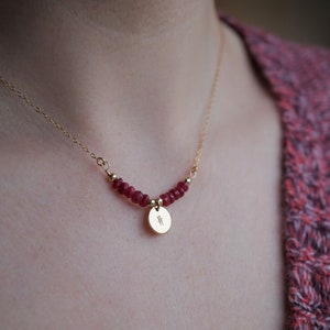 Puede incluir: Un collar de cadena de oro con un pequeño colgante de disco de oro y una fila de cuentas de piedras preciosas rojas.