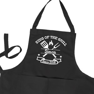 May include: Black cotton apron with a white design that says "King of the Grill" with a grill spatula and tongs crossed over a flame. The design also includes the name "Simon" and the text "Since 2018".