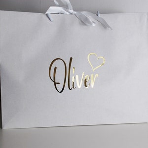 May include: White gift bag with a gold foil design. The bag has a white ribbon and the name "Oliver" with a heart above it.