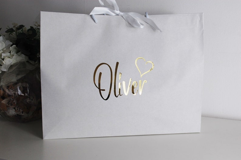 May include: White gift bag with a gold foil design. The bag has a white ribbon and the name "Oliver" with a heart above it.