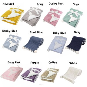 May include: Twelve knitted baby blankets in various colors, including mustard, grey, dusky pink, sage, dusky blue, steel blue, baby blue, navy, baby pink, purple, coffee, and white. Each blanket has a white pom-pom on one corner and a white satin ribbon tied in a bow on the top.