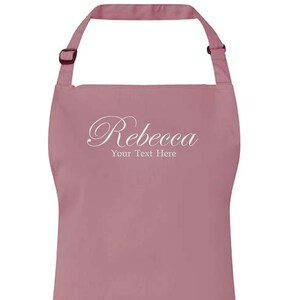 Op de afbeelding: Een roze schort met de naam "Rebecca" en de tekst "Your Text Here" in wit cursief schrift.
