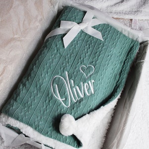 May include: A soft, green knitted baby blanket with a white pom-pom and a white embroidered heart and the name "Oliver".
