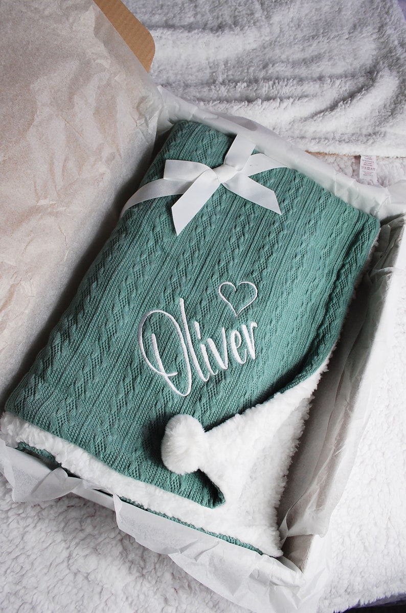 May include: A soft, green knitted baby blanket with a white pom-pom and a white embroidered heart and the name "Oliver".