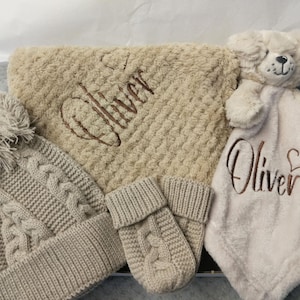 May include: A beige knitted hat with a pom-pom, a pair of beige knitted mittens, a beige plush blanket with the name "Oliver" embroidered on it, and a white plush security blanket with the name "Oliver" and a heart embroidered on it.