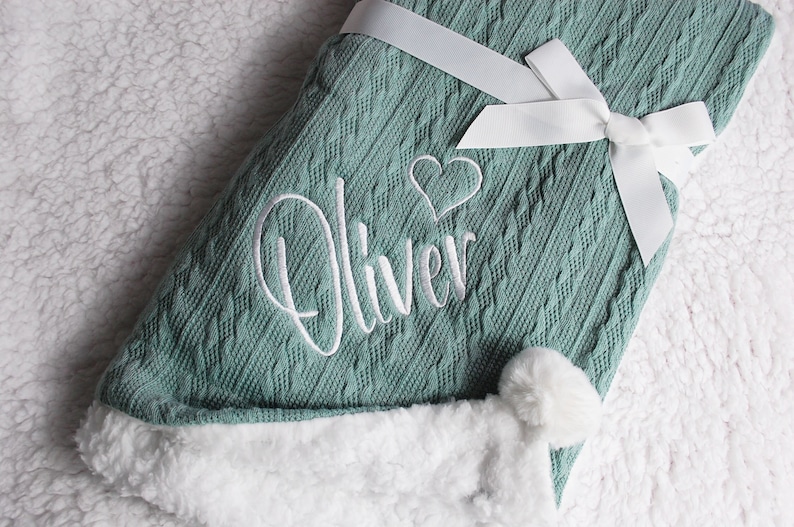 May include: A soft, green knitted baby blanket with a white pom-pom trim and a white satin ribbon tied in a bow. The blanket is personalized with the name "Oliver" and a small heart embroidered in white.