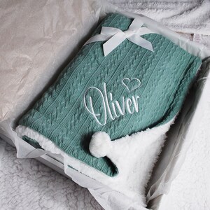May include: A soft, green knitted baby blanket with a white pom-pom and white embroidery that says "Oliver" with a heart.