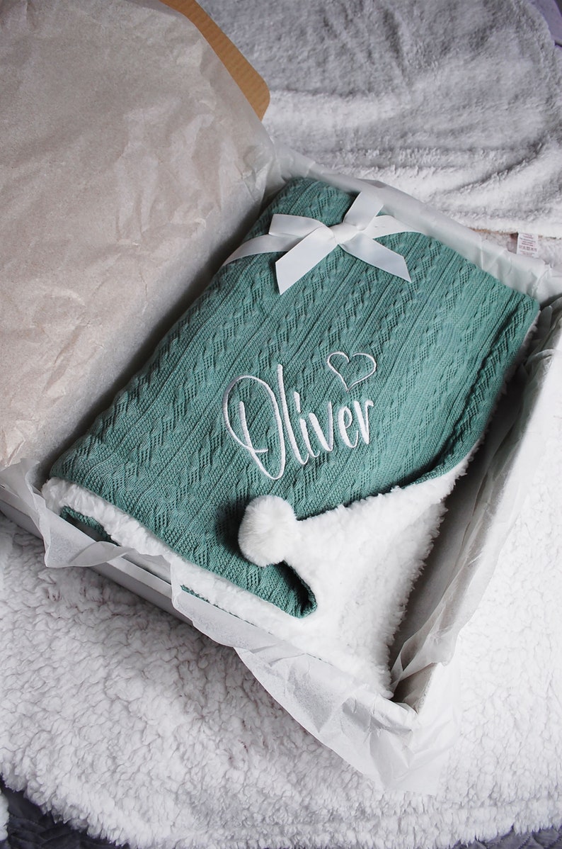 May include: A soft, green knitted baby blanket with a white pom-pom and white embroidery that says "Oliver" with a heart.