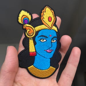 May include: Embroidered patch of a blue-skinned Hindu deity with a yellow crown and a peacock feather headdress. The deity has a red dot on their forehead and is wearing a gold necklace.