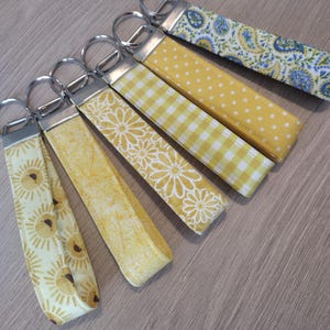 May include: A collection of fabric keychains with various patterns, including yellow polka dots, gingham, floral, and paisley designs. Each keychain has a silver metal ring and a fabric loop. The keychains are arranged on a light wood surface.