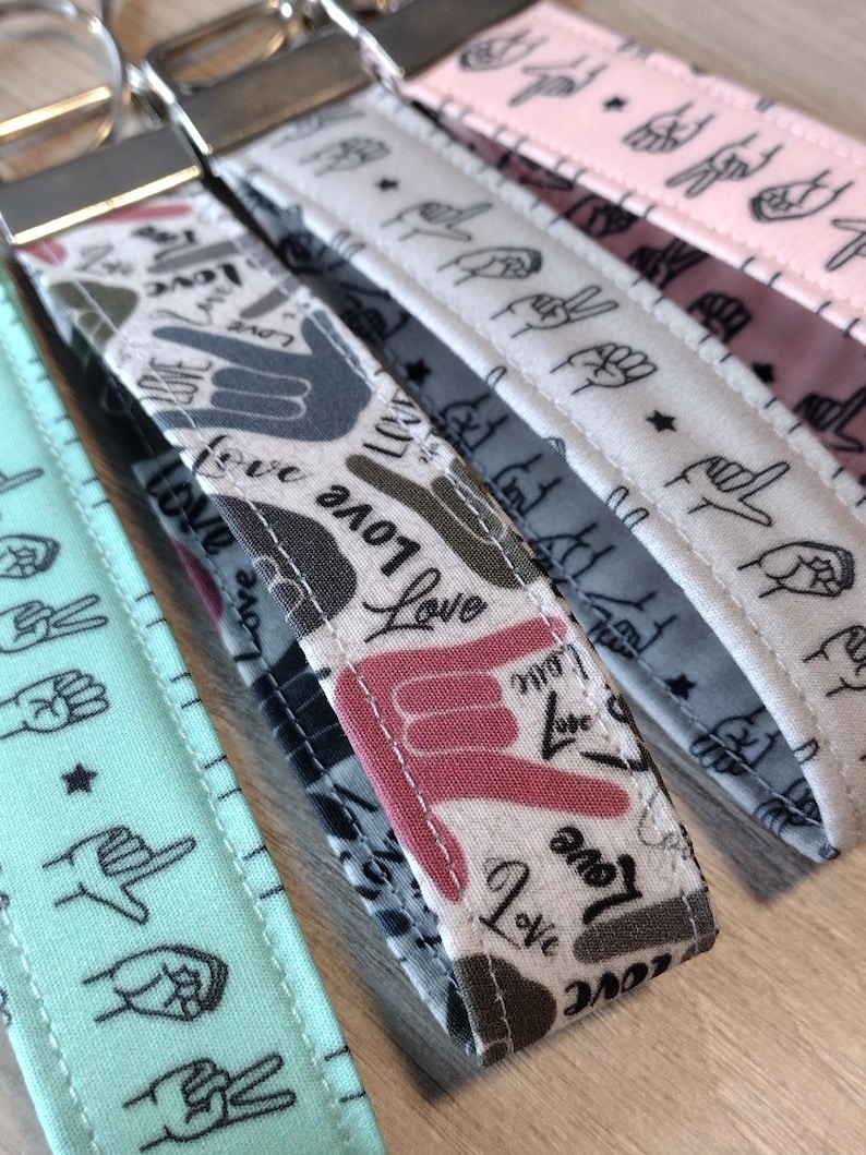 May include: Four keychains with different patterns. The keychains are made of fabric and feature a variety of hand signs and the word "love" in different fonts. The keychains are all attached to silver key rings.