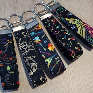 May include: Five black keychains with different colorful musical designs. The designs include musical notes, guitars, and treble clefs. The keychains are made of fabric and have silver key rings.