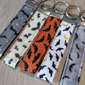 May include: Five different fabric keychains with a bat pattern. The keychains are in gray, cream, orange, white, and gray. Each keychain has a silver metal key ring.