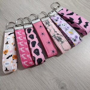 May include: A collection of seven fabric wristlet keychains featuring various Halloween-themed patterns in shades of pink, black, and white. The designs include pumpkins, ghosts, bats, coffins, spiders, skeletons, and floral motifs with the text BOO.