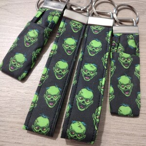 May include: Four black and green keychains with a repeating pattern of green zombie faces. Each keychain has a silver metal ring and a silver metal clasp.