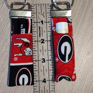 May include: Two fabric lip balm holders with key rings. One holder is empty, the other contains a lip balm. The holders are red, black, and white, featuring a bulldog and the word "Georgia". The ruler shows the holders are approximately 4 inches long.