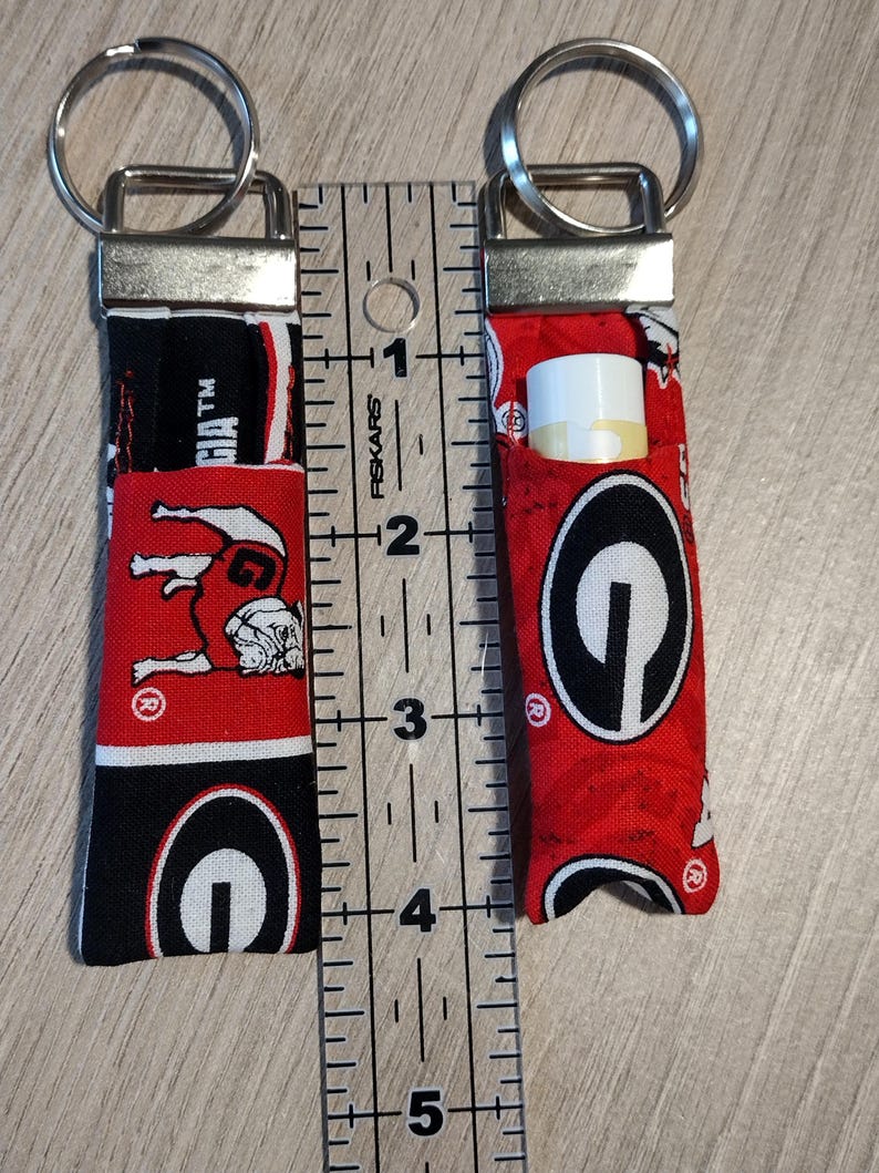May include: Two fabric lip balm holders with key rings. One holder is empty, the other contains a lip balm. The holders are red, black, and white, featuring a bulldog and the word "Georgia". The ruler shows the holders are approximately 4 inches long.